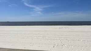 Une plage de sable blanc parsemée d’empreintes, un ciel bleu dégagé et une eau calme.