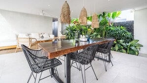 A modern dining area with a wooden table and black wicker chairs, surrounded by lush green plants and a stone wall. - Green Flow Villa 20 (Ubud)