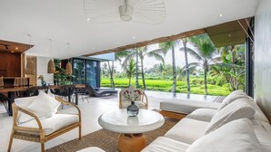 A modern living room with a large window, white sofas, a round coffee table, and a dining area with a wooden table and chairs. - Green Flow Villa 20 (Ubud)