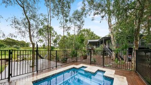 A swimming pool with a wooden deck and metal railing, surrounded by trees and a house. - Fig Tree Guest House (Pokolbin)