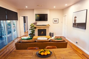 A modern living room with a fireplace, a television mounted above it, and a dining area with a wooden table and leather sofas.