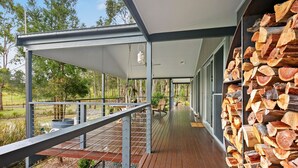 A covered outdoor deck with a wooden floor, metal railings, and a stack of firewood. - Fig Tree Guest House (Pokolbin)