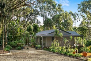 A single-story house with a dark roof, surrounded by dense vegetation and trees.