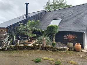 Une maison avec un toit en pente, une cheminée, un mur en pierre et diverses plantes en pot.
