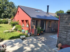 A red barn with a sloped roof, a wooden shed, a concrete patio, and a lawn with a sun lounger and potted plants. - Maison Atypique (Quéven)