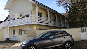 Un immeuble de deux étages avec un balcon, une voiture garée devant et un portail.