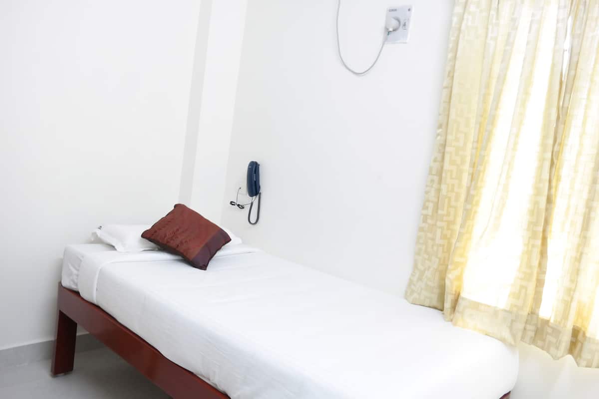 A single bed with white bedding and a brown pillow in a room with a plain white wall, a telephone, and a patterned curtain.