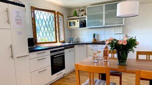 Une cuisine moderne avec des meubles blancs, un four encastré, une table à manger avec des chaises et une fenêtre donnant sur un paysage verdoyant.