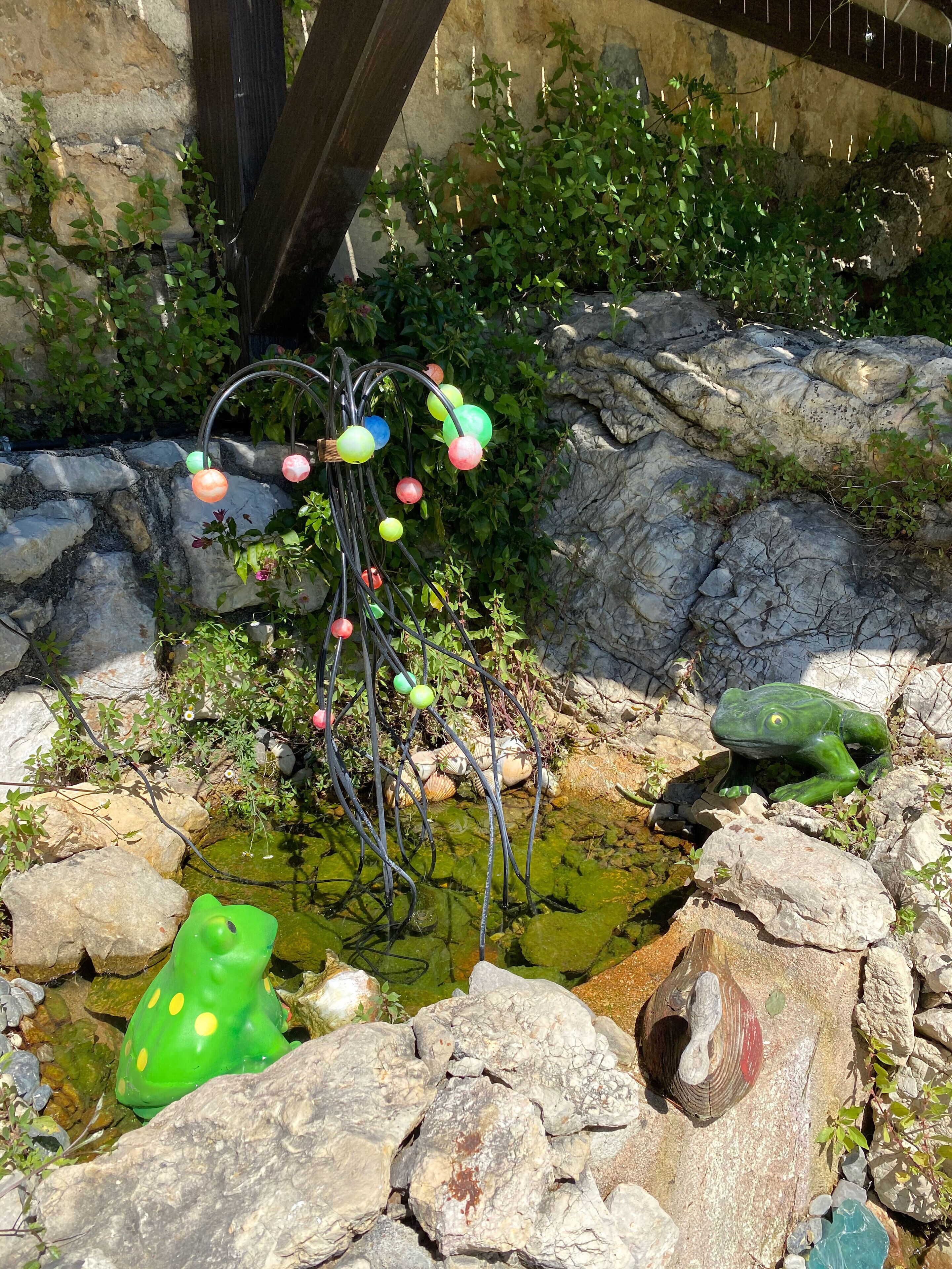 Ein Teich mit Steinen und einem Frosch, umgeben von Grünpflanzen und einer dekorativen Leuchte.