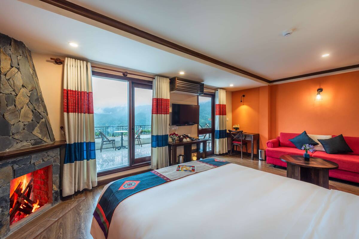 Ein Hotelzimmer mit Kamin aus Stein, einem Bett mit farbenfroher Tagesdecke, einem roten Sofa, einem kleinen Tisch und einem Balkon mit Tisch und Stühlen.