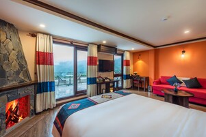 Habitación de hotel con chimenea de piedra, una cama con una colcha de colores, un sofá rojo, una pequeña mesa y un balcón con mesa y sillas.
