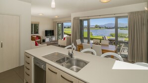 A modern kitchen with a large island, a dining area with a view of a lake, and a cozy seating area with a TV. - Great Matarangi Location Lakeside Gem overlooking the golf course! (Matarangi)