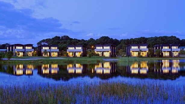 A row of modern, multi-story buildings with large windows, reflecting in a calm body of water, surrounded by greenery and trees. - Great Matarangi Location Lakeside Gem overlooking the golf course! (Matarangi)