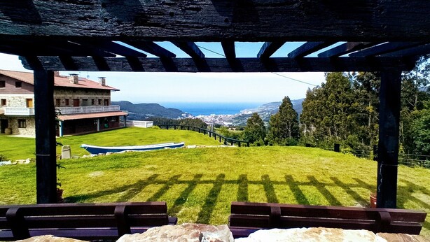 Una pérgola de madera con bancos que da a un césped con vistas a un pueblo y montañas al fondo.