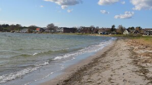 Een strand met een zandrijk strand en golven, huizen op de achtergrond en een heldere lucht.