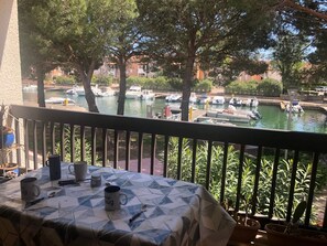 A balcony with a table set for two, overlooking a marina with boats and a building in the background. - Saint Cyprien Appart 4 Personnes Lagune (Saint-Cyprien)