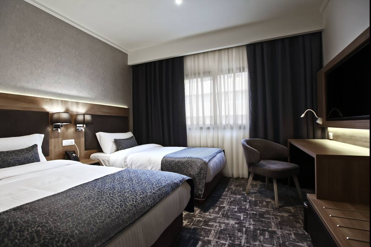 A hotel room with two beds, a desk, a chair, a TV, and a window with curtains.
