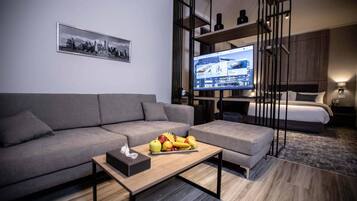 A modern hotel room with a grey sofa, a wooden coffee table with fruits, a television, and a bed with a headboard.