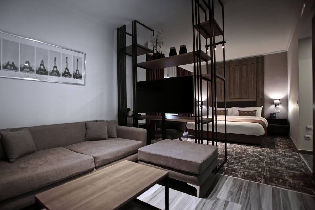 A modern hotel room with a sofa, a coffee table, a TV, a bed, and a nightstand.
