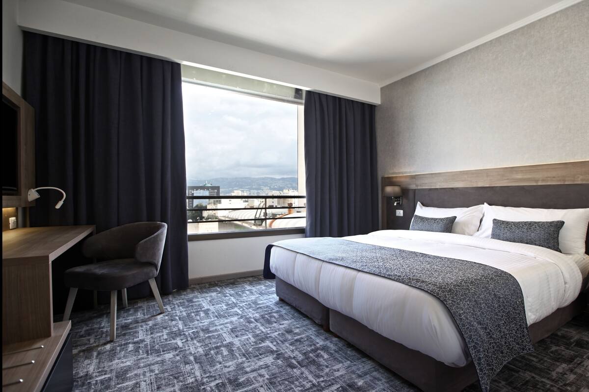 A hotel room with a large bed, a desk, a chair, and a view of the city through the window.