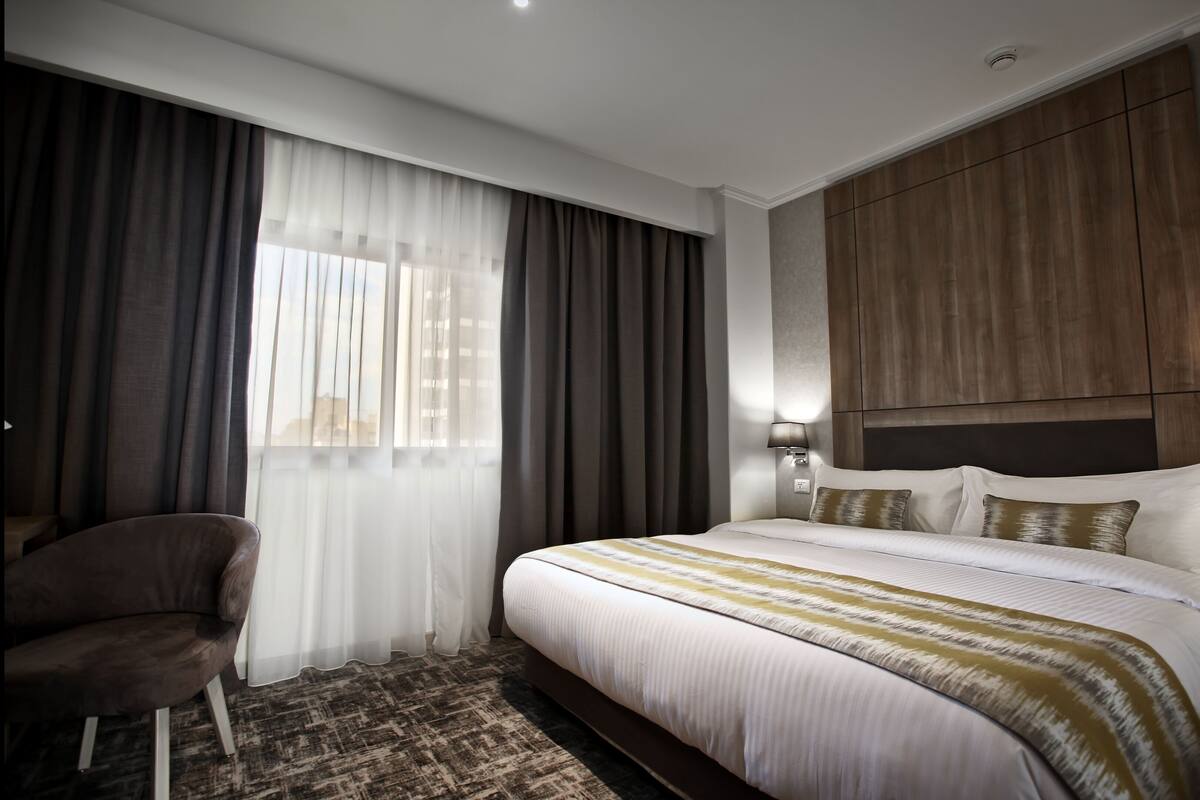 A modern hotel room with a large bed, a chair, and a view of the city through sheer curtains.