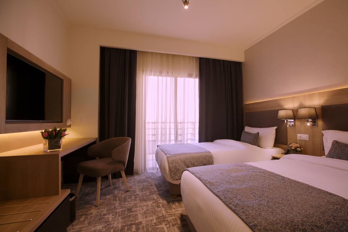 A hotel room with two beds, a desk, a chair, a TV, and a window with curtains.