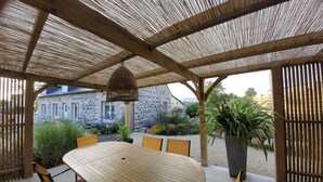 Ein überdachter Essbereich im Freien mit Holztisch und -stühlen, einer hängenden Leuchte und Blick auf ein Steinhaus im Hintergrund.