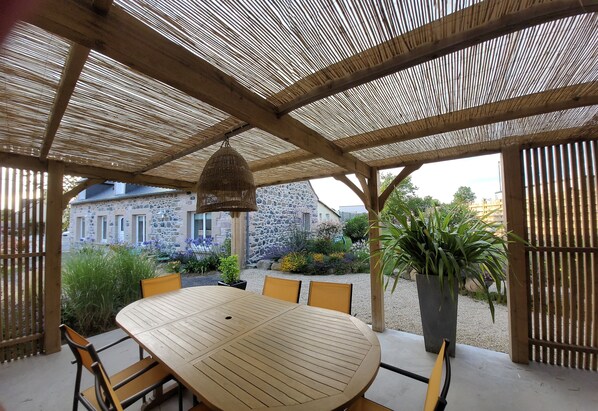 A covered outdoor dining area with a wooden table and chairs, a hanging light fixture, and a view of a stone building in the background. - Comfortable house, enclosed garden, all inclusive, linen, housework. 5 mn from the beach (Plurien)