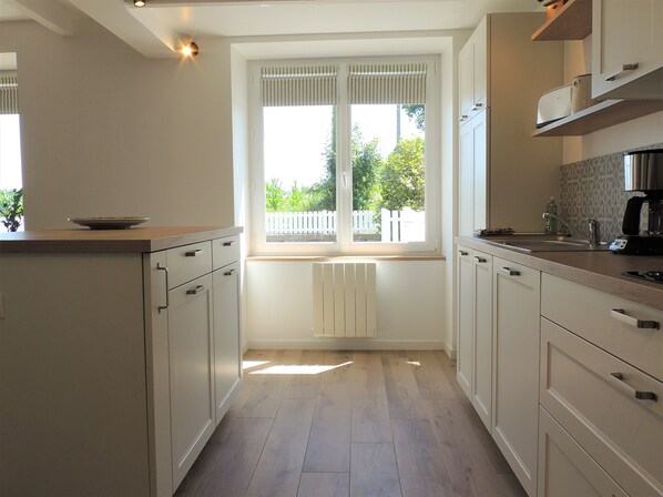 A kitchen with white cabinets, a window with blinds, a radiator, and a coffee maker on the counter. - Comfortable house, enclosed garden, all inclusive, linen, housework. 5 mn from the beach (Plurien)
