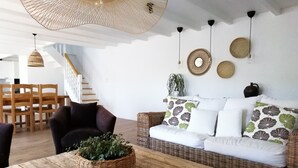 Ein gemütliches Wohnzimmer mit einem Rattan-Sofa, einem Holztisch mit einer Pflanze und einem Essbereich mit Holzsesseln und einer Treppe.