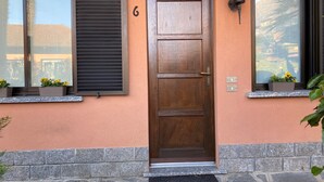 Eine Holztür mit der Aufschrift '6' darüber, begleitet von Fenstern mit Fensterläden und Blumentöpfen.