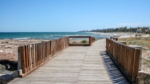 En träpromenad som leder till en strand med fri utsikt över havet och avlägsna byggnader.