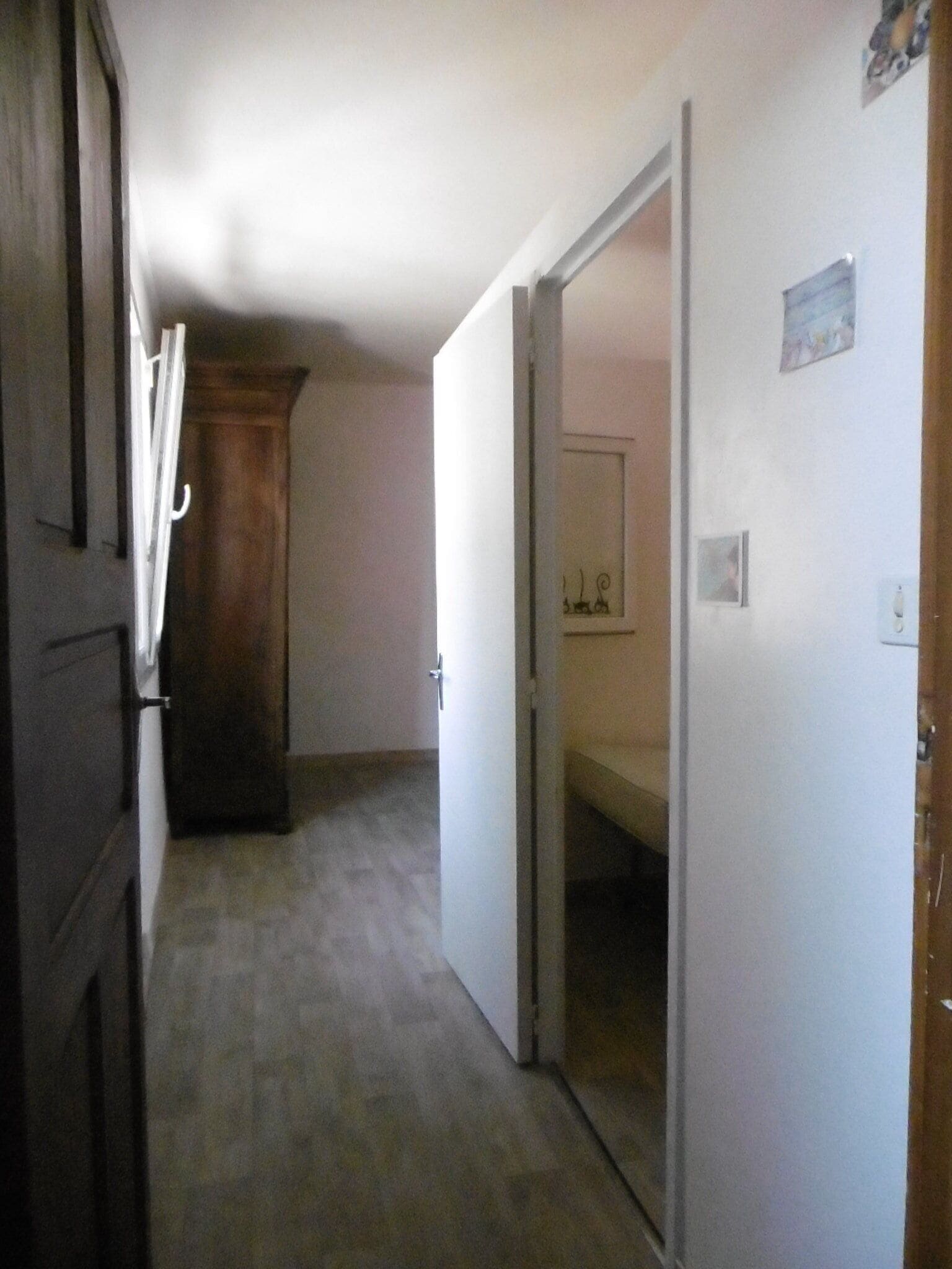 A narrow hallway with a wooden door on the left, a white door at the end, and a bed visible through an open door on the right.
