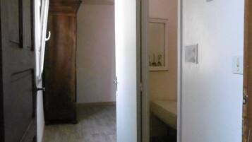 A narrow hallway with a wooden door on the left, a white door at the end, and a bed visible through an open door on the right.