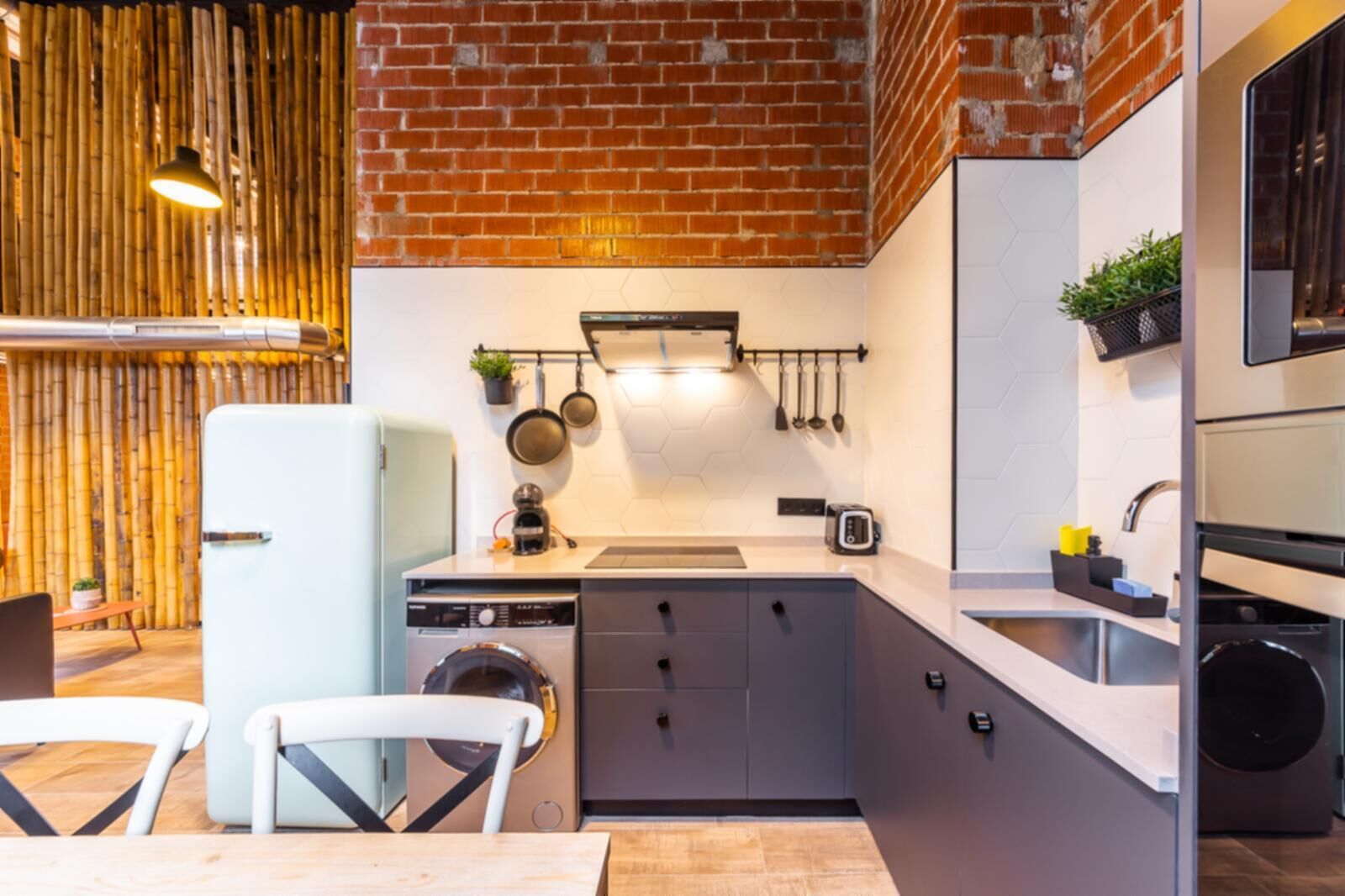 A compact kitchen with a brick wall, a washing machine, a sink, and a dining area with a table and chairs.