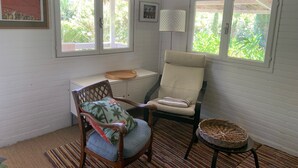 Ett rum med vita väggar, två fönster, en trästol med en kudde, ett litet bord med en flätad korg och en inramad tavla på väggen.