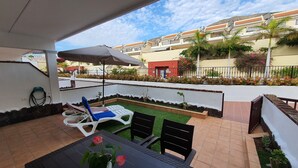 A patio with a table, chairs, and an umbrella. There is a small garden area with grass and plants.