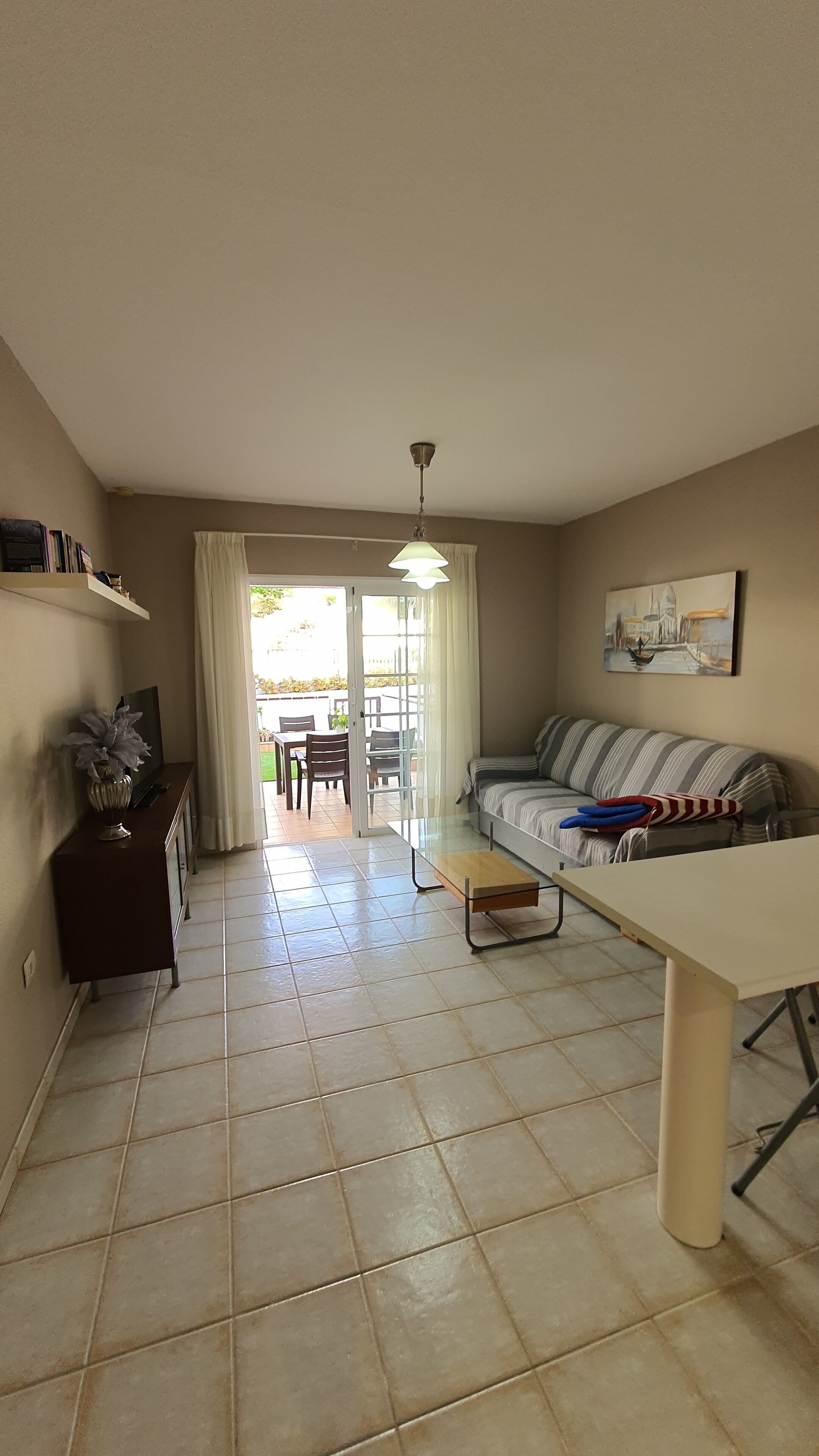 Un salon comprenant un canapé, un téléviseur, une table et une bibliothèque. Une porte donne accès à un patio aménagé avec une table et des chaises.
