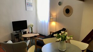 Ein gemütliches Wohnzimmer mit Flachbildfernseher, einem kleinen runden Tisch mit Stühlen, einer Blumenvase und einer Wanduhr.