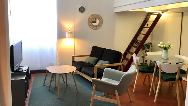 Ein Wohnzimmer mit einer Couch, einem Couchtisch, einem Fernseher, einer Lampe, einem kleinen Tisch mit einer Vase und einer an der Wand stehenden Leiter.
