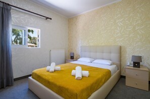 Una cama bien tendida con un edredón amarillo y toallas enrolladas, una mesita de noche con una lámpara y una ventana con cortinas.