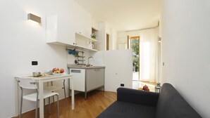 A compact living space with a kitchenette, a dining area with a white table and chairs, a dark-colored sofa, and a view of the outdoors through a door. - Sicily 2 - 3 - 4 Person With Pool (Altavilla Milicia)