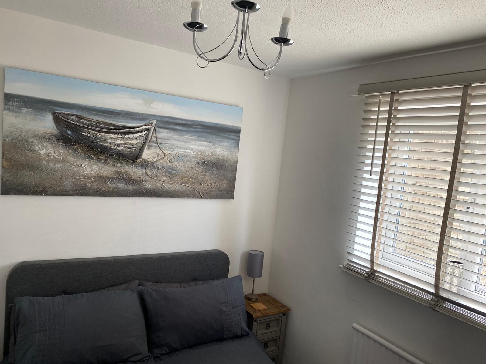 A bedroom with a bed, a painting of a boat on water, a chandelier, and a window with blinds.