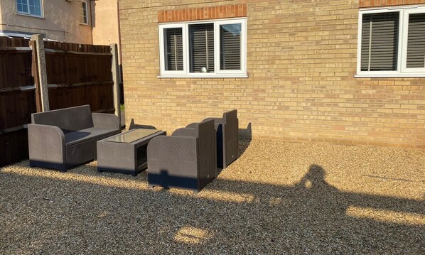 An outdoor seating area with a table and chairs, a wooden fence, and a brick building with windows.