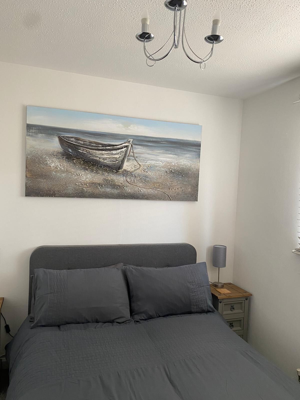 A bedroom with a bed, a nightstand, a lamp, and a painting of a boat on a beach.