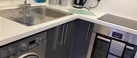 A compact kitchen with a stainless steel sink, a built-in washing machine, a coffee maker, and a microwave.