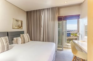 Habitación de hotel con cama, escritorio, silla, ventana con cortinas y un cuadro en la pared.