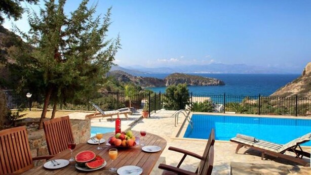 Un espace repas aménagé au bord de la piscine, avec une table dressée pour deux personnes, offrant une vue sur une mer d’un bleu limpide et un paysage montagneux.