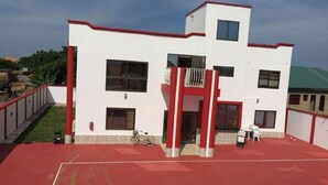 A two-story building with a red and white exterior, a balcony, and a red sports court in front. - Rasta Tees Place - Standard (Adenta)