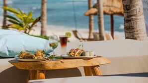 Una mesa de madera con un plato de comida, una bebida y un recipiente pequeño, frente a un paisaje de playa con palmeras y una estructura de techo de paja.